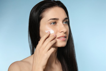 Portrait of beautiful young woman applying facial cream on color background