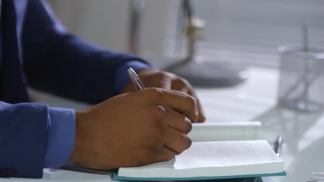 Tracking close up shot of hands of unrecognizable black man sitting at office desk and using pen and notebook for planning