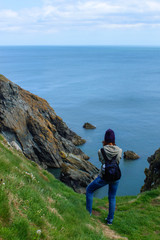 Naklejka na ściany i meble Unrecognizable young female tourist enjoying the view after hiking in Howth, Ireland. Amazing Irish landscape with green rugged cliffs rising from the sea.