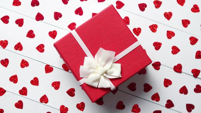 Valentine's Day decoration composition. Boxed gift placed on heart shaped red sequins on white wooden table. Romantic background. Flat lay, top view.