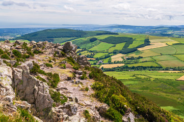 Fototapeta na wymiar Mountain peak landscape with rugged rocks and distant hills covered in different shades of green. View from the top of The Great Sugar Loaf Mountain in Wicklow, Ireland on a sunny Summer day.