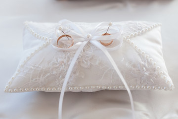 A pair of wedding rings on a white pillow. In the center of the pillow is a ribbon bow. Close-up