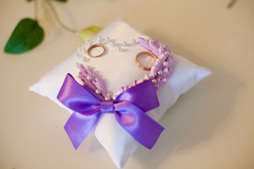 A pair of wedding rings on a white pillow. In the middle of the pillow, a lilac heart-shaped ornament is made.
