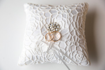 A pair of wedding rings on a white pillow. In the center of the pillow is a brilliant decoration.