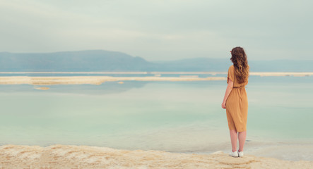 Back view of stylish girl wearing dress on seaside, Dead Sea beach. Travel, summer vacation, holiday, freedom concept. Digital detox. Sea background with copy space. Banner.