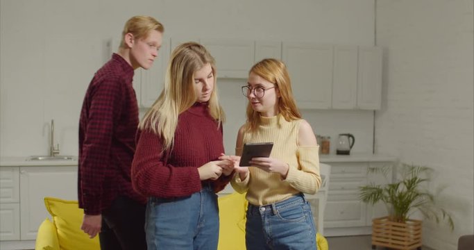 Cheerful female friends chatting and networking with digital tablet in domestic room. Positive group of teenagers with tablet pc discussing social media content and threads online indoors.