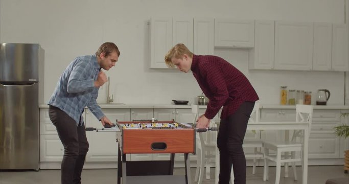 Cheerful excited best male frineds playing table football while enjoying freetime together in loft apartment. Positive roommates having fun and relaxing playing table soccer after work at home.