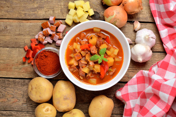 View from above of potato and sausage goulash, pepper, onion and garlic on wooden table and red towel