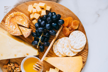 Flat lay with various types of cheese, grapes, nuts, honey and cracker in wooden board on marble