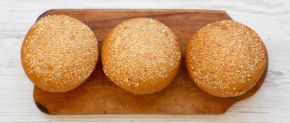 Fresh burger buns with sesame seeds on wooden board on white wooden surface, overhead view. Fast...