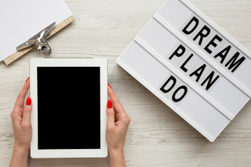 Female hands hold tablet with blank screen, modern board with text 'Dream plan do', clipboard over white wooden background, overhead view. From above, flat lay, top view.
