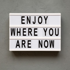 Naklejka premium 'Enjoy where you are now' words on lightbox over concrete background, top view. Flat lay, from above, overhead.