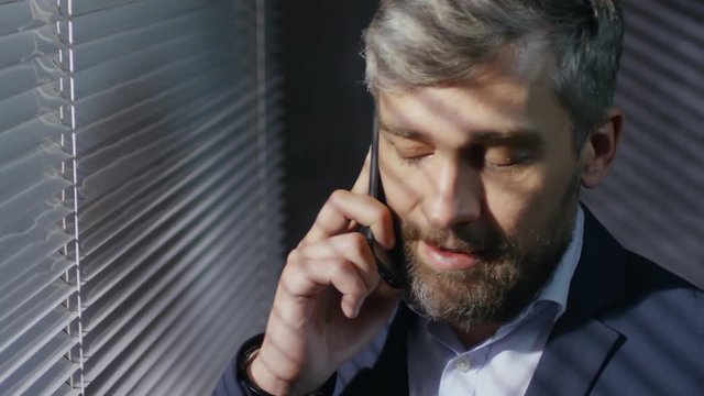 Close up face of bearded office worker looking out of office window with slatted venetian blinds on it while discussing project on phone