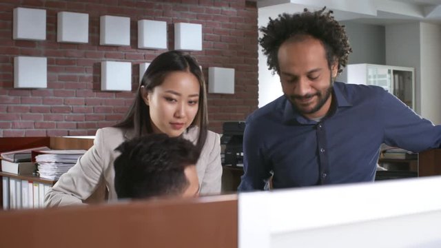Group of three multi ethnic colleagues discussing work in office, one of them sitting at desk and the other two standing near him