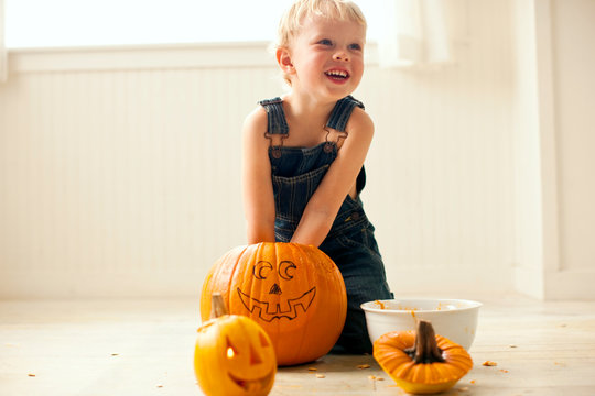 Young boy looks up and laughs as he kneels on a floor and digs both hands into a big pumpkin with a face drawn on it with a bowl and a finished small Jack O'Lantern with a candle lit inside in front of him.