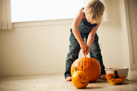 Young boy bends over to use both hands to hold a tool to scrape out the insides of a big pumpkin with a face drawn on it to make it into a Jack O'Lantern like the small one sitting in front of him.