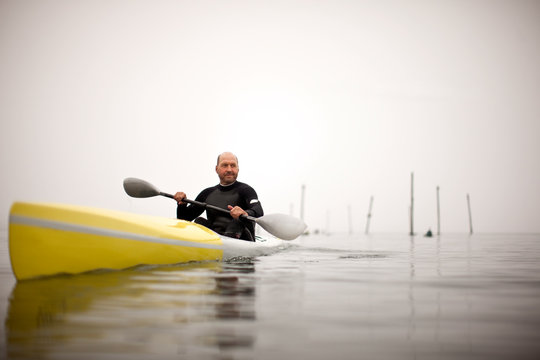 Male kayaker smiles for a portrait as he paddles in the waters of a foggy harbour.