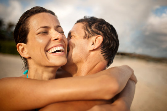 Smiling mid-adult woman being kissed on the cheek by her husband on a beach.