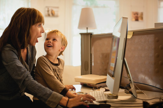 Young boy smiles up at his mother and she makes a surprised face as they sit together at a home study desk in front of a large desktop computer screen.
