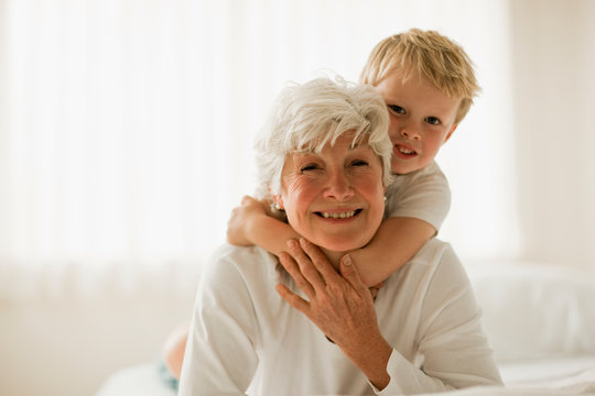 Young boy lies on his grandmother's back with his arms around her neck as they lie on their fronts on a bed and smile for a portrait.