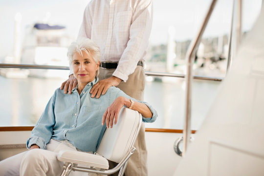 Senior womans sits on a boat deck while her husband stands with his hands on her shoulders.
