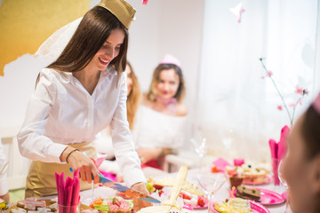 The smiling young bride stewardess standing near the festive table at the hen party and cutting a cake