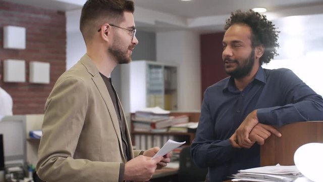 Medium shot of two friendly young men of different ethnicities talking to each other at work in office while colleagues working in the background