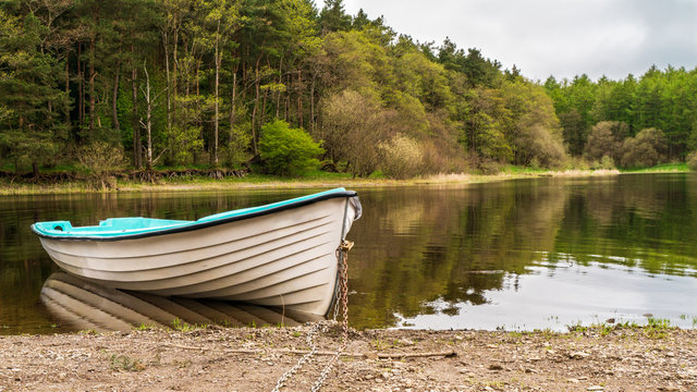 Peaceful morning landscape with a white fishing boat tied and anchored at Blessington Lake shore in Dublin, Ireland, on a beautiful forest background with trees reflected in the water.