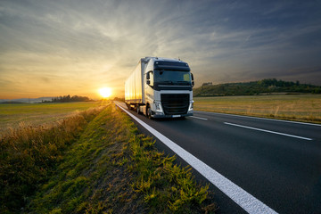 White truck arriving on the asphalt road in rural landscape at sunset