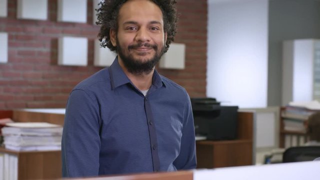 Portrait of friendly young Middle Eastern man standing in modern office and smiling at camera while colleagues working in the background