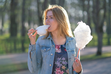 charming caucasian girl walks in the spring Park in a denim jacket and eats pink cotton candy,...