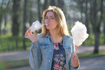 charming caucasian girl walks in the spring Park in a denim jacket and eats pink cotton candy,...