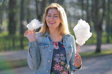 charming caucasian girl walks in the spring Park in a denim jacket and eats pink cotton candy,...
