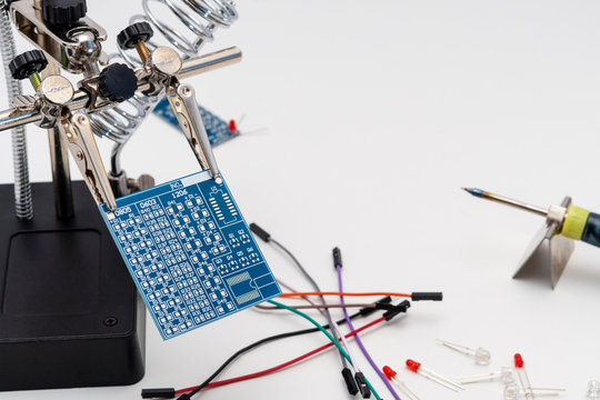 Blue circuit board in soldering helping hands (third hand or auxiliary clip tool) before technological soldering process with LEDs (Light-emitting diode), wires and soldering iron on the background.