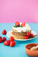 pancakes in a plate with whipped cream, honey, strawberries, mint and sweet cherries on a bright blue and pink background.