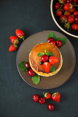 pancakes in a plate with strawberries, mint, honey and cherry on a dark black background.