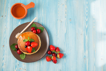 pancakes in a plate with strawberries, mint, honey and cherry on a bright blue background.