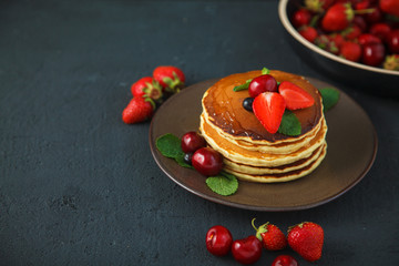 pancakes in a plate with strawberries, mint, honey and cherry on a dark black background.