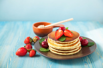 pancakes in a plate with strawberries, mint, honey and cherry on a bright blue background.