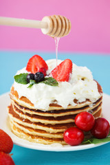 pancakes in a plate with whipped cream, honey, strawberries, mint and sweet cherries on a bright blue and pink background.
