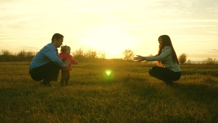 baby goes on lawn from dad to mom. child takes first steps in park. mom and dad play with kid on grass at sunset. family happiness concept