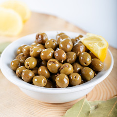 Green olives in bowl with leaves of and lemon on wooden background. Green olives from Antakya, Turkey.