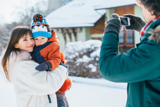 Caucasian father takes pictures on phone of happy mother and little baby toddler son in her arms in snowy winter day outdoor. Family together concept.