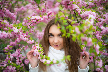 Fototapeta premium .A beautiful fair-haired girl with green eyes in a white coat and a white scarf stands in the forest in a pink cherry blossom and holds branches with her hands. Horizontal photography.
