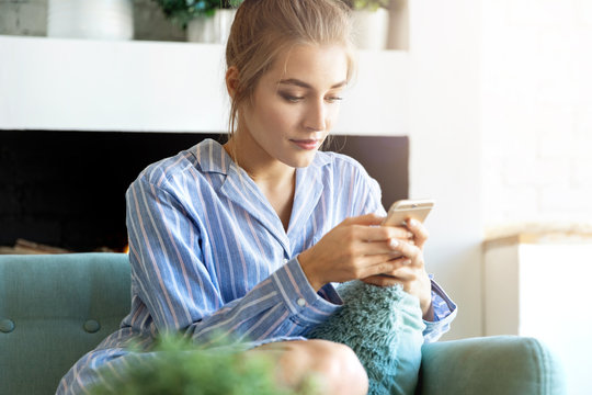 Portrait of sweet caucasian model texting on trendy smartphone. Pretty girl sitting in pyjamas at home with nice interior. Technology and morning concept. Blurred background