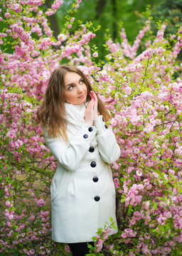 A beautiful fair-haired girl in a white coat is posing and dreaming in the garden of cherry blossoms. Vertical photography