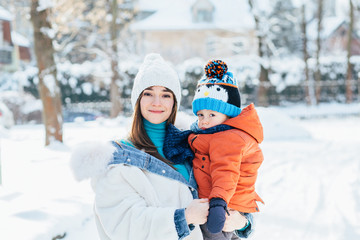Cheerful woman in white jacket holding her sad little toddler son on her arms outdoor in winter day. Holidays, people, family, fun and vacation concept.