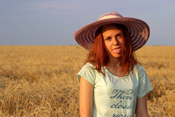 Girl in a hat showing tongue on yellow wheat field in summer. Funny girl