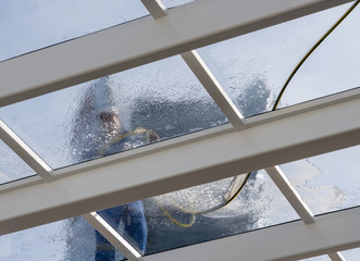 Male worker washing large expanse of glass roof over swimming pool with hose pipe