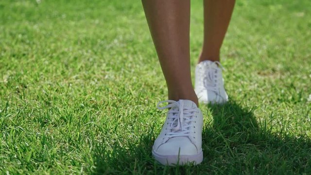Female feet in white sneakers, are walking along the bright green grass. Walking on the lawn in white shoes: close-up, only legs. A shadow on the grass. Slender female legs in white shoes